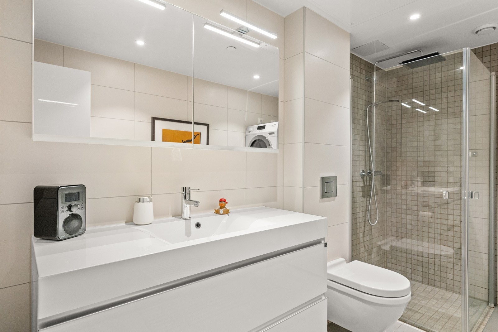 Praktisk dusjhjørne med innfellbare glassdører og regnfallsdusj, servantskap med skuffer i høyglans hvit utførelse og nedsenket servant i kompositt materiale. Galleribilde