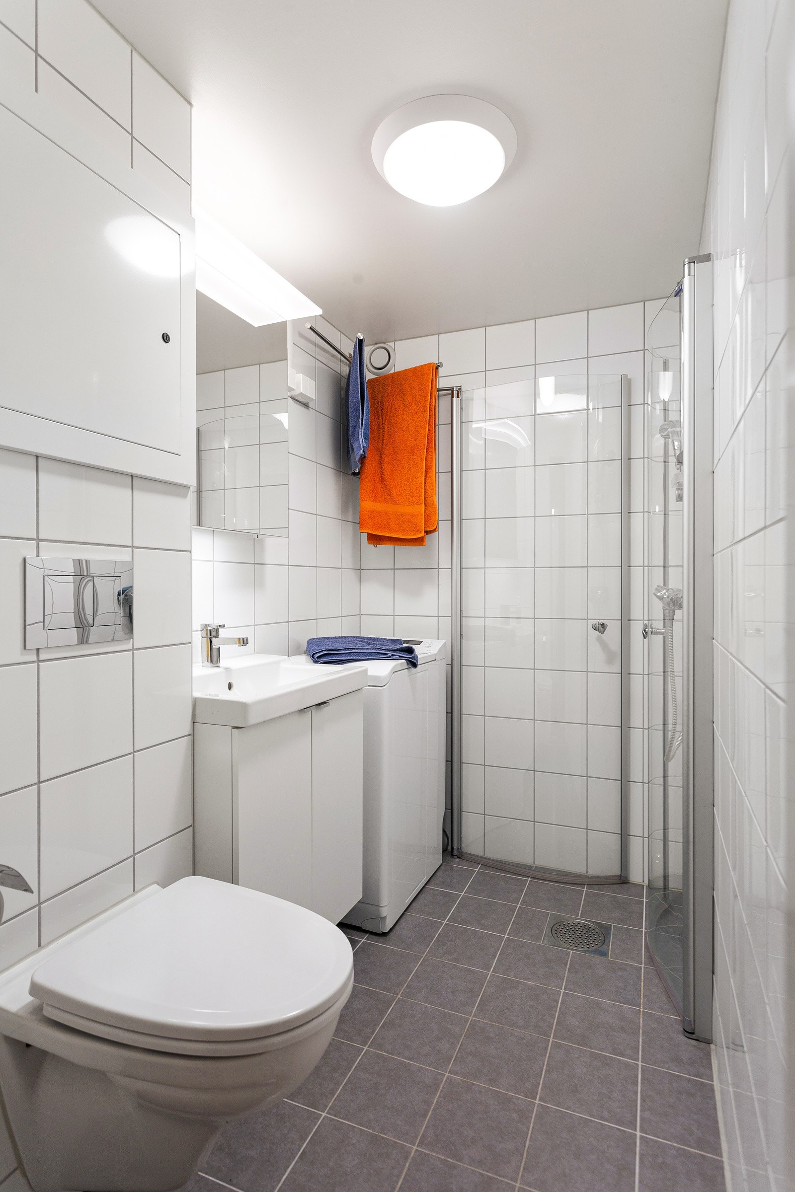 Baderomsinnredningen består av dusjhjørne med to buede fronter i herdet glass, servant i skapinnredning, veggmontert klosett med innfliset sisterne og opplegg for vaskemaskin Galleribilde