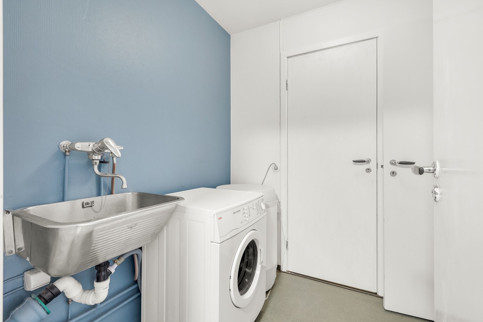 Vaskerommet har grått belegg på gulvet, mens veggene er malt i hvitt og en dus blåfarge. I rommet er det skyllekum i rustfritt stål og opplegg for vaskemaskin. Galleribilde