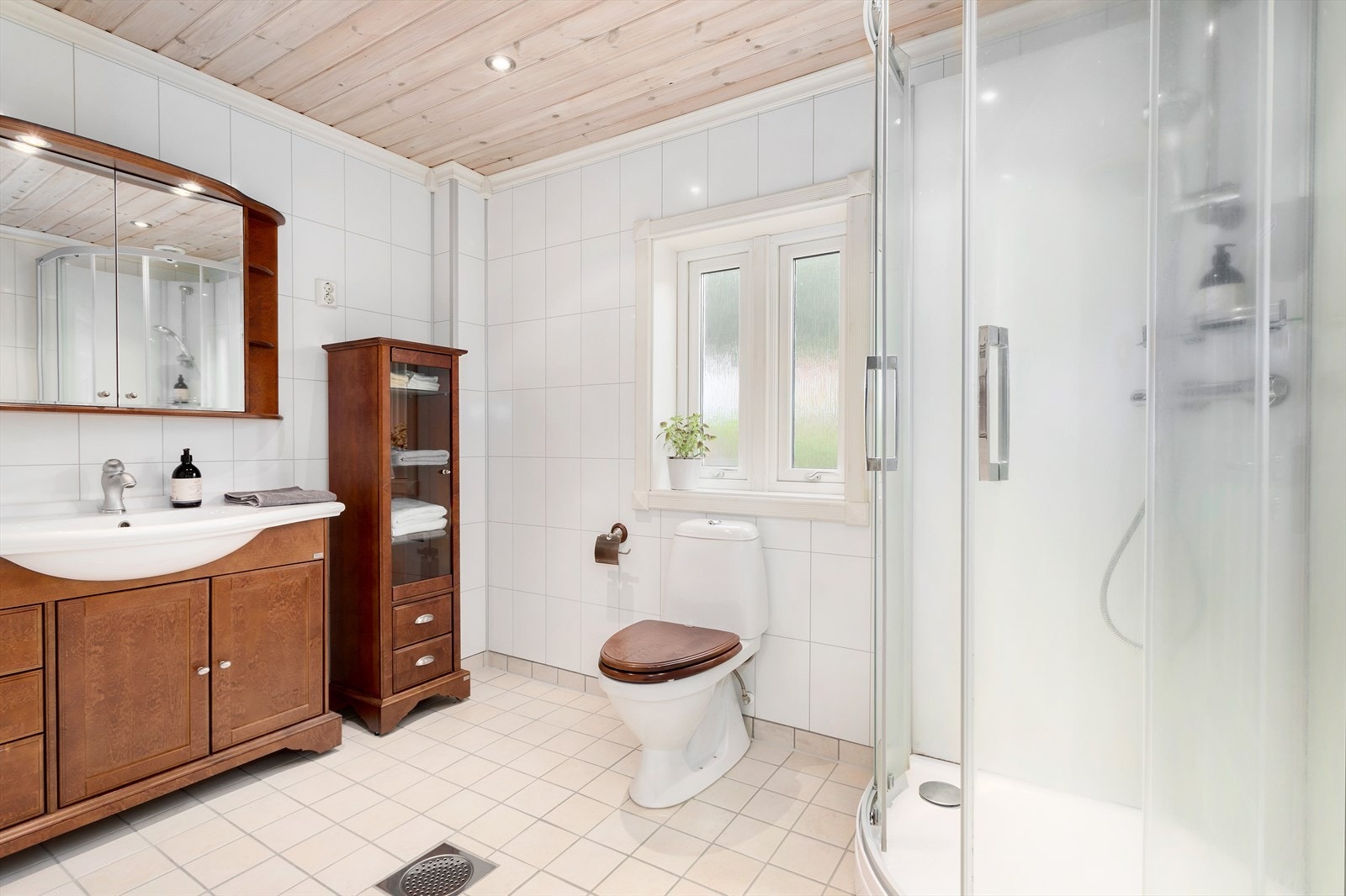 Romslig, flislagt bad som er innredet med toalett, dusjkabinett og servant på baderomsinnredning. Galleribilde