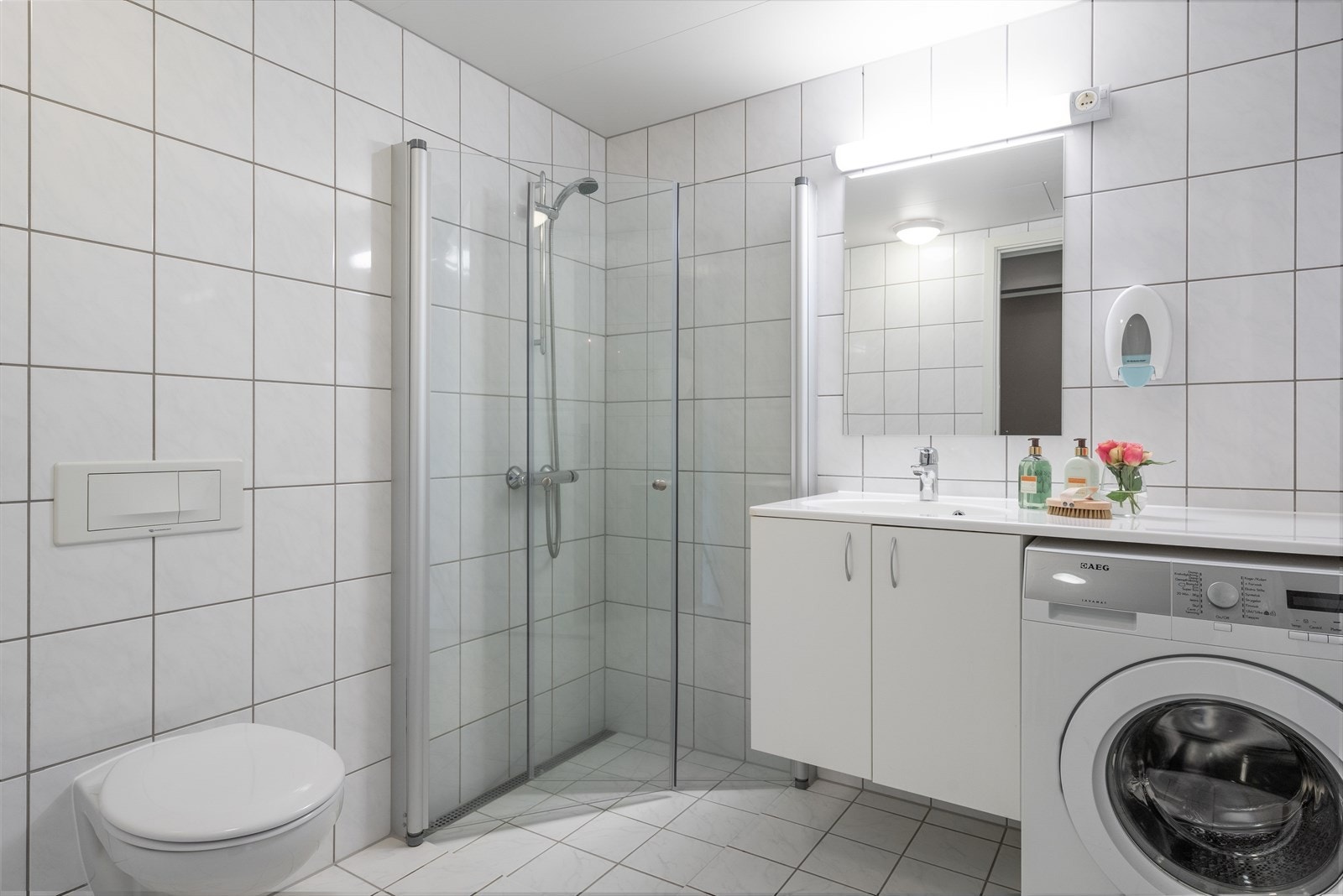 Bad med flislagte overflater, baderomsinnredning, dusj med glassdører og vegghengt toalett. Opplegg for vaskemaskin. Galleribilde