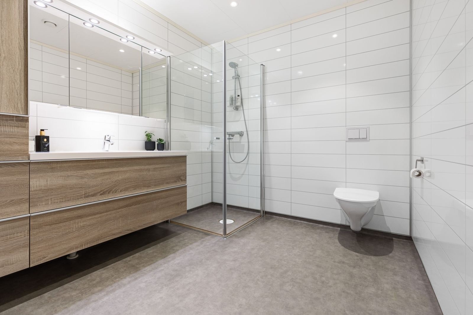 Badet er utstyrt med dusjhjørne, innfellbare dusjdører, mekanisk avtrekk, servant, servantskap og veggmontert wc Galleribilde