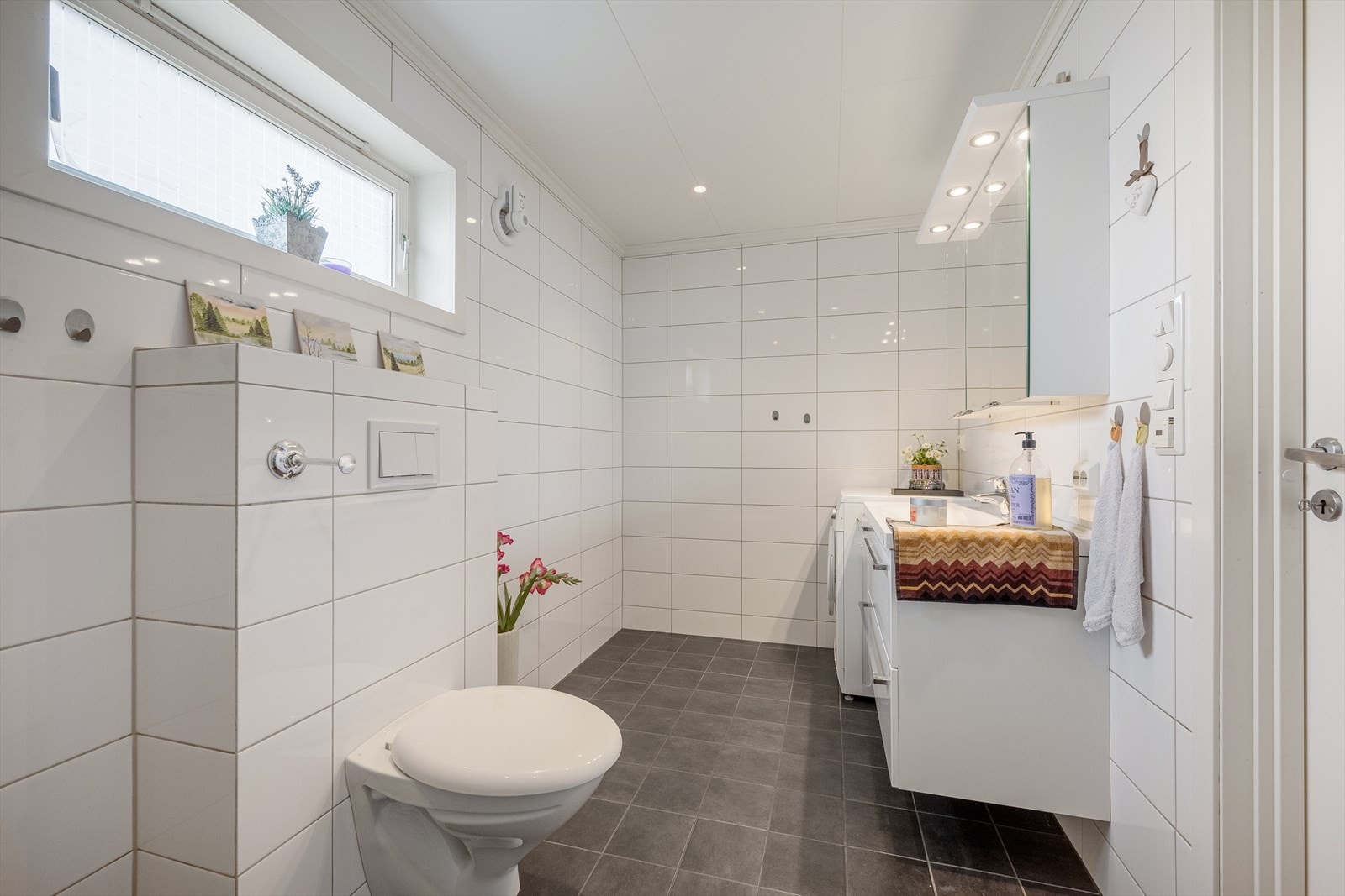 Badet er innredet med dusj, vegghengt toalett og servant med skuffeseksjon og speil. Galleribilde