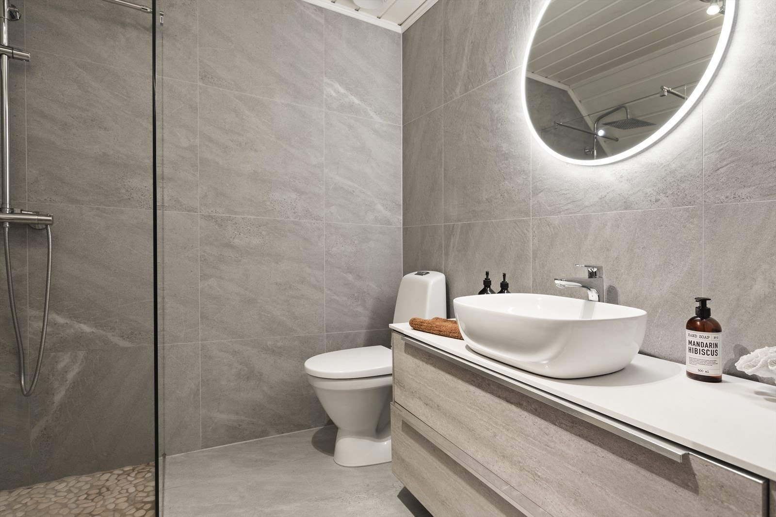 Baderomsinnredning av servant på benk med innredning av to skuffer og lysspeil. Videre er det sokkeltoalett og dusjnisje med rullestein, regnfallsdusj og glassvegg. Galleribilde