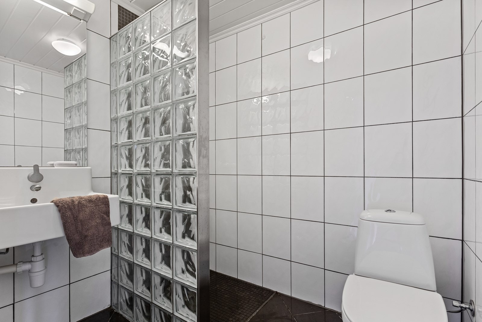 Badet har flislagte overflater og er utstyrt med toalett, dusj med glassbyggerstein og servant. Galleribilde
