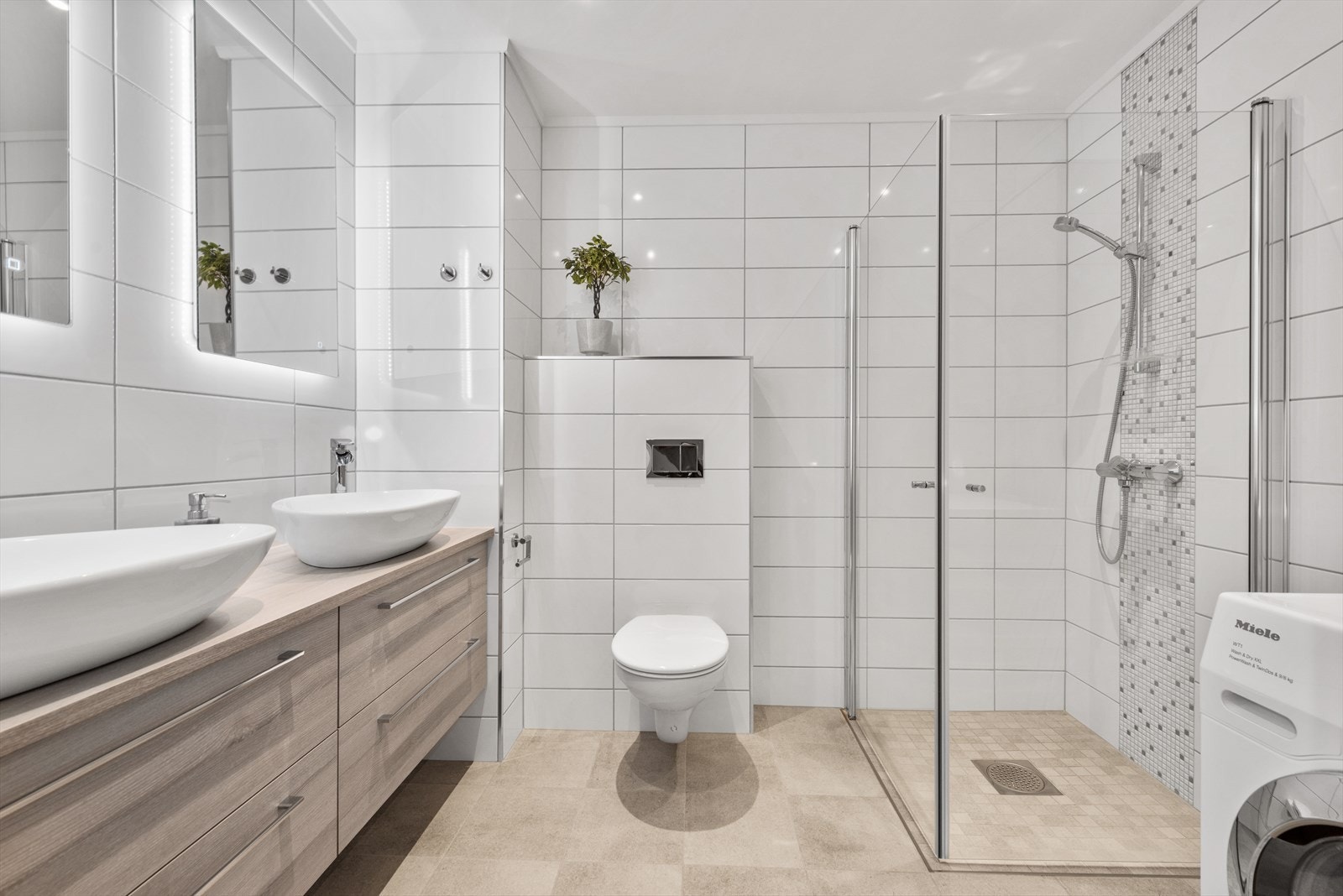 Flislagt baderom utstyrt med vegghengt klosett, dusjhjørne med glassdører og innredning med to servanter. Galleribilde