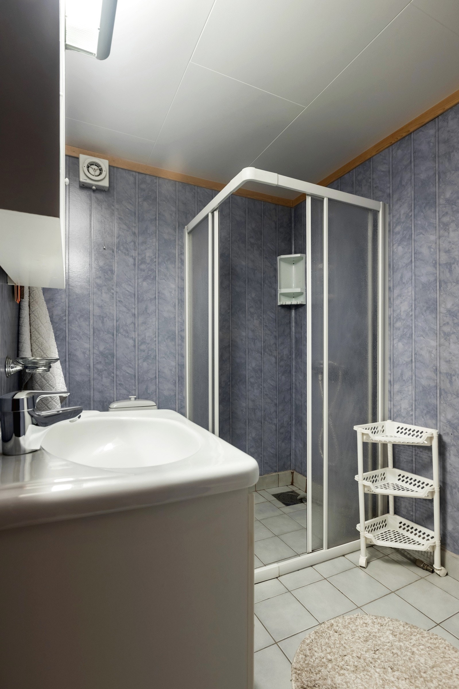 Bad innehar flislagt gulv, våtromstapet på vegger og takess-plater i himling i lyse farger. Badet er innredet med klosett, dusjhjørne, servant med servantskap, overskap med speil og overlys. Galleribilde