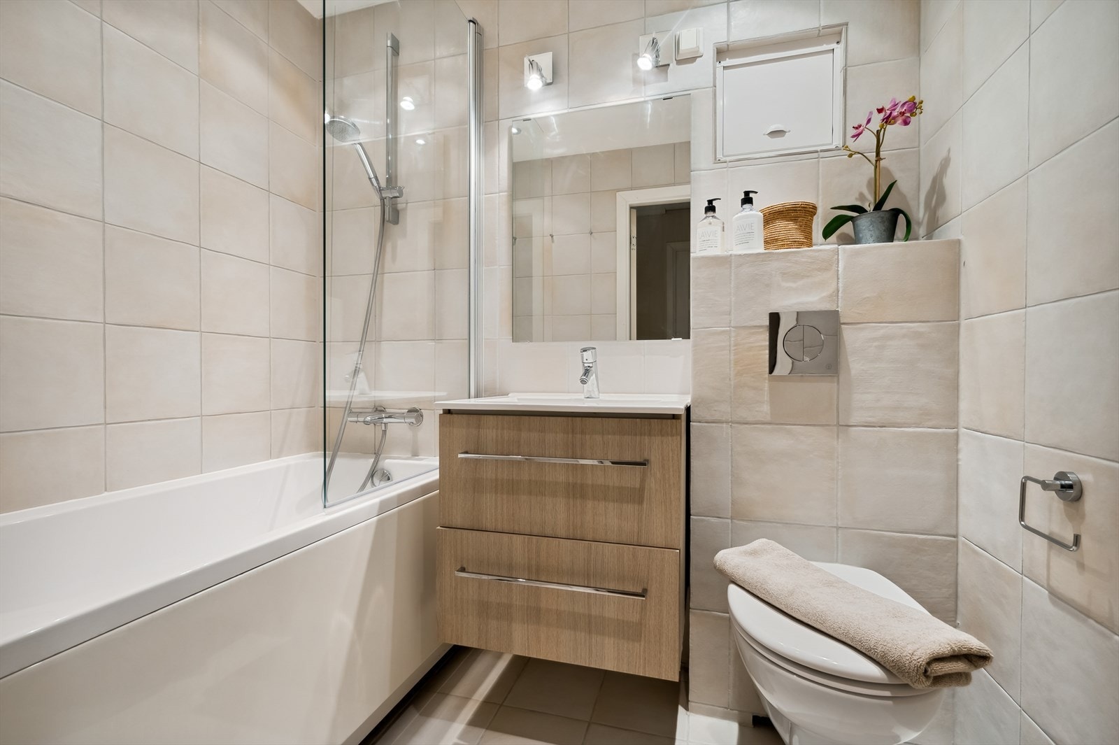 Flislagt baderom utstyrt med veggfestet klosett, dusj i badekar med dør i herdet glass og servant med tilhørende benkeskap. Galleribilde