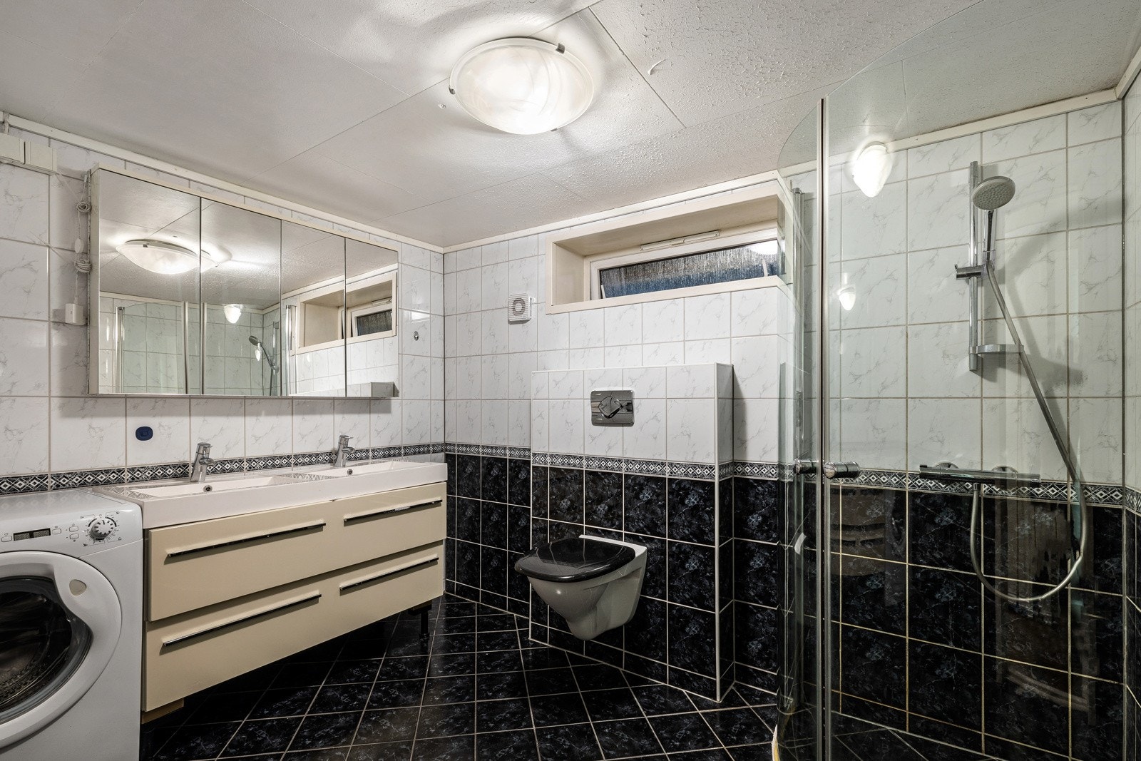 Badet i hybelen er av flislagte overflater med vegghengt toalett, benkeskap med servant, dusjdører og opplegg for vaskemaskin. Galleribilde
