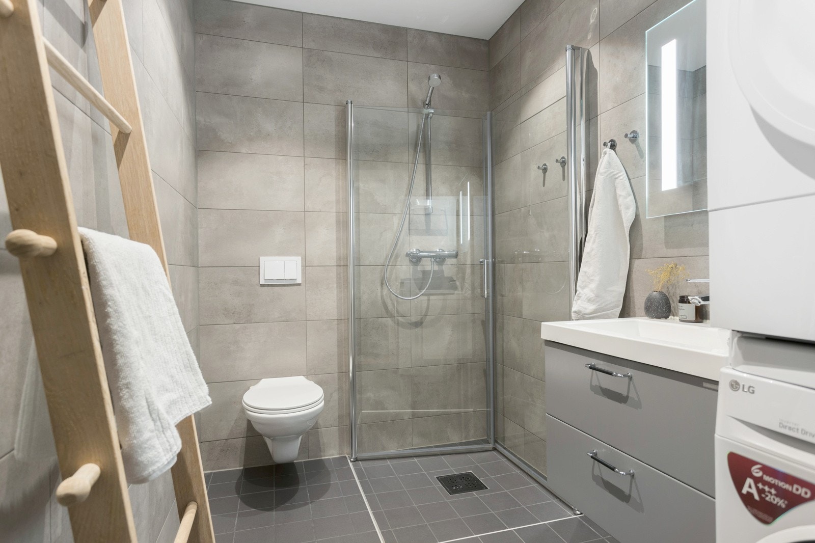 Bad med veggmontert toalett, dusjdører i herdet glass, opplegg for vaskemaskin/tørketrommel, veggspeil med integrert belysning, baderomsinnredning med malte glatte fronter og heldekkende servant. Galleribilde