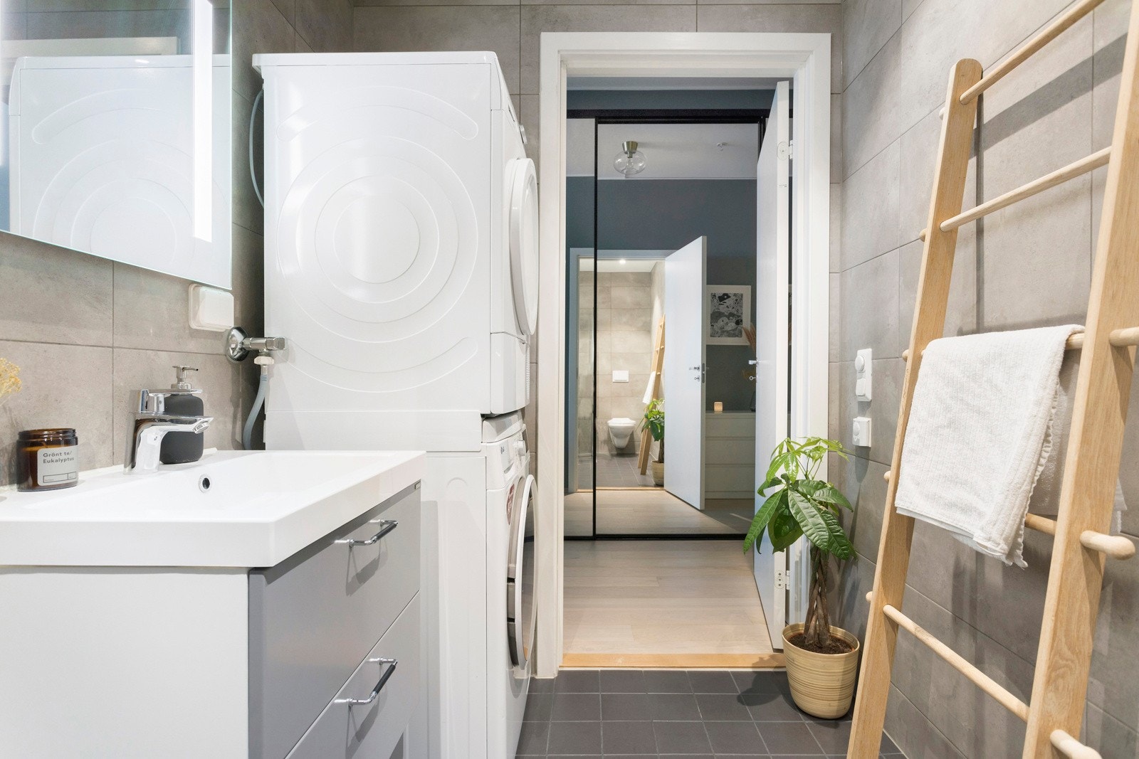 Bad/vaskerom med veggmontert toalett, dusjdører i herdet glass, opplegg for vaskemaskin/tørketrommel, veggspeil med integrert belysning, baderomsinnredning med malte glatte fronter og heldekkende servant. Flislagte overflater. Galleribilde