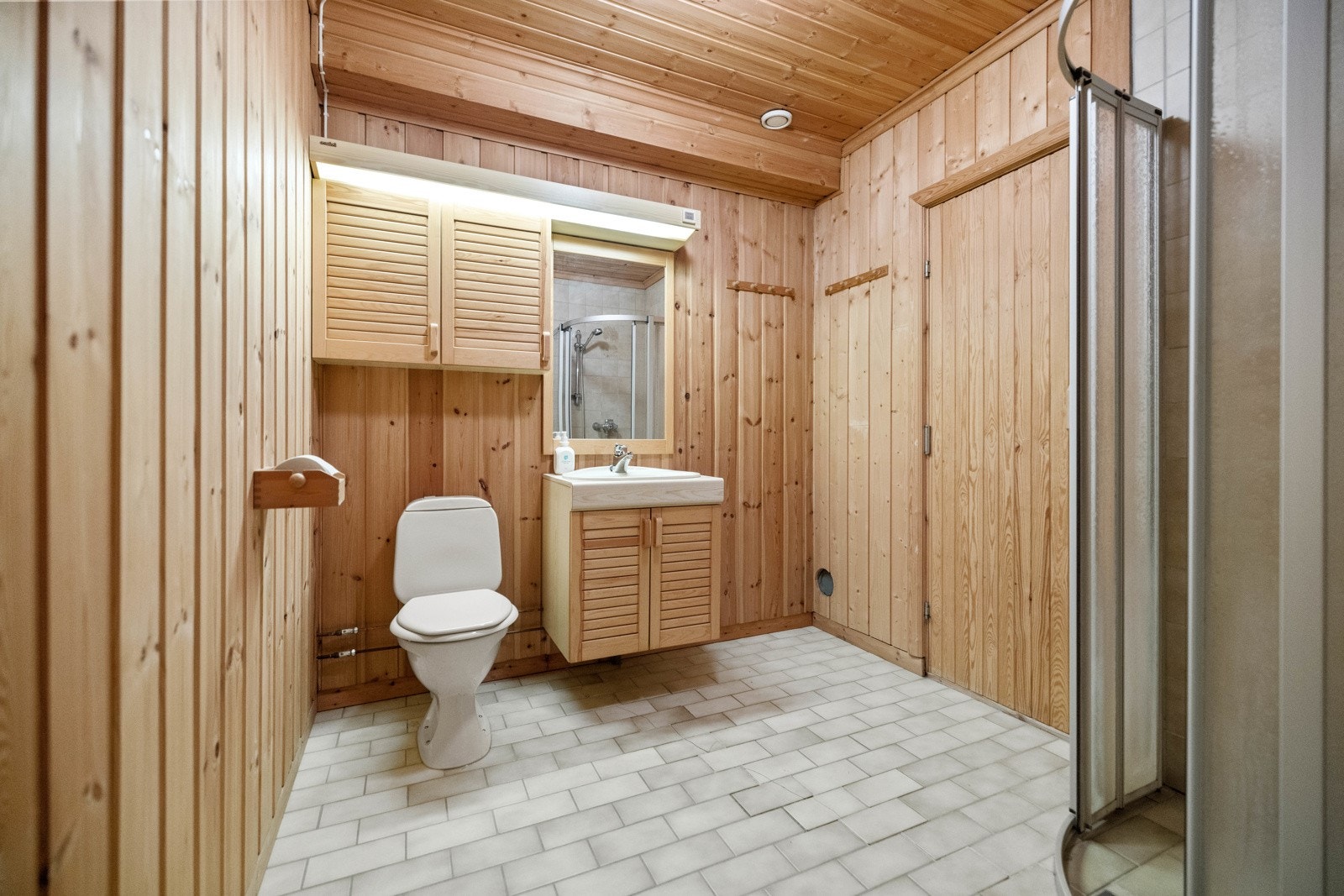 Bad i underetasjen med inngang til badstue. Fliser på gulv med varmekabler. Vegger av delvis flis og panel. Servant med servantskap, toalett, dusjhjørne med styrenglass vegger og dører. Galleribilde