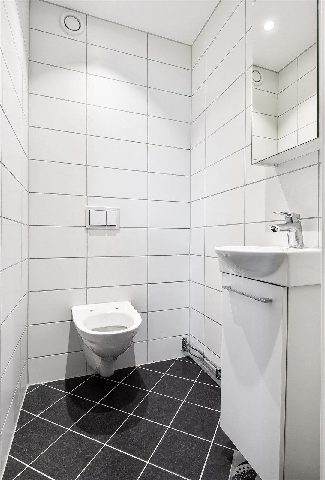 Gjestetoalett fra 2021 med flislagt gulv og vegger. Platehimling med downlights, sluk med synlig klemring og vannlås. Servant med underskap og speilskap over vask. Galleribilde