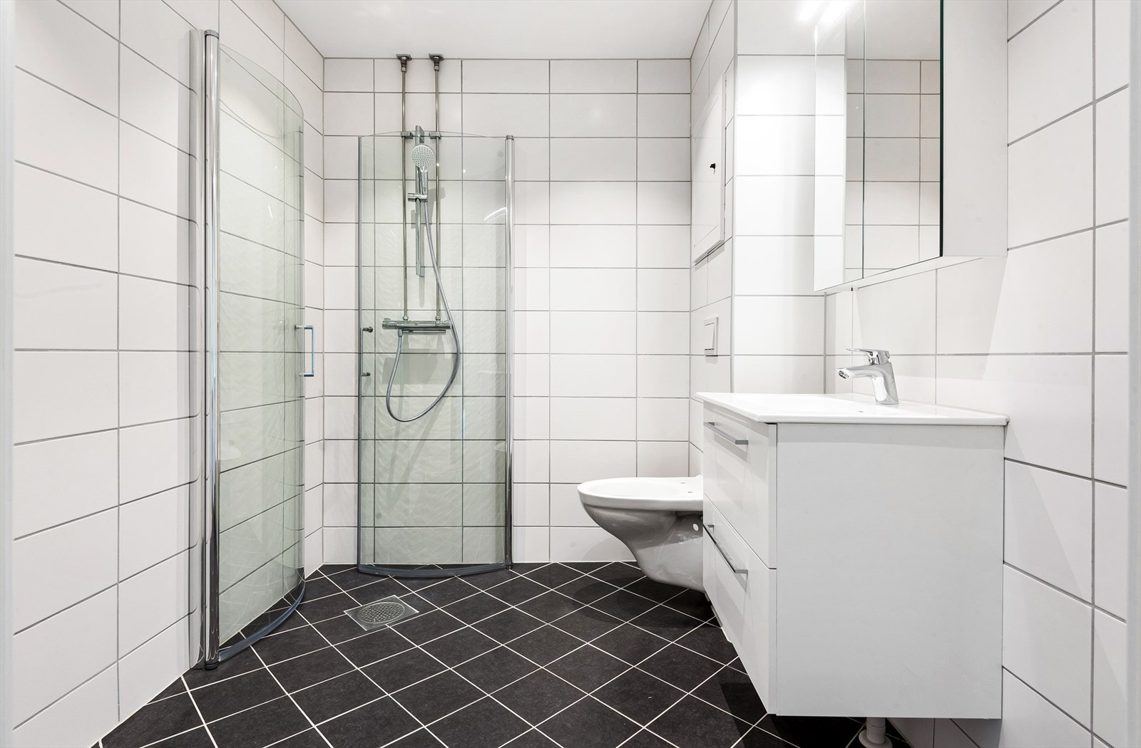 Baderom fra 2021 med dusjhjørne med dører i herdet glass, servant med underskuffer, speilskap og lys over servant.
Vegghengt toalett og opplegg for vaskemaskin / tørketrommel. Galleribilde