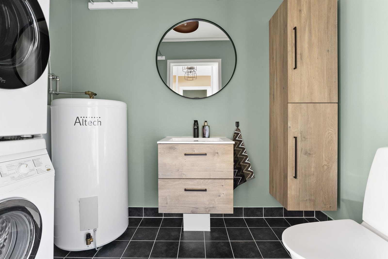 Vaskerom med flislagt gulv og grønnmalte overflater. Her er det innredet med toalett, servant med skap og oppbevaringsskap på vegg. Galleribilde