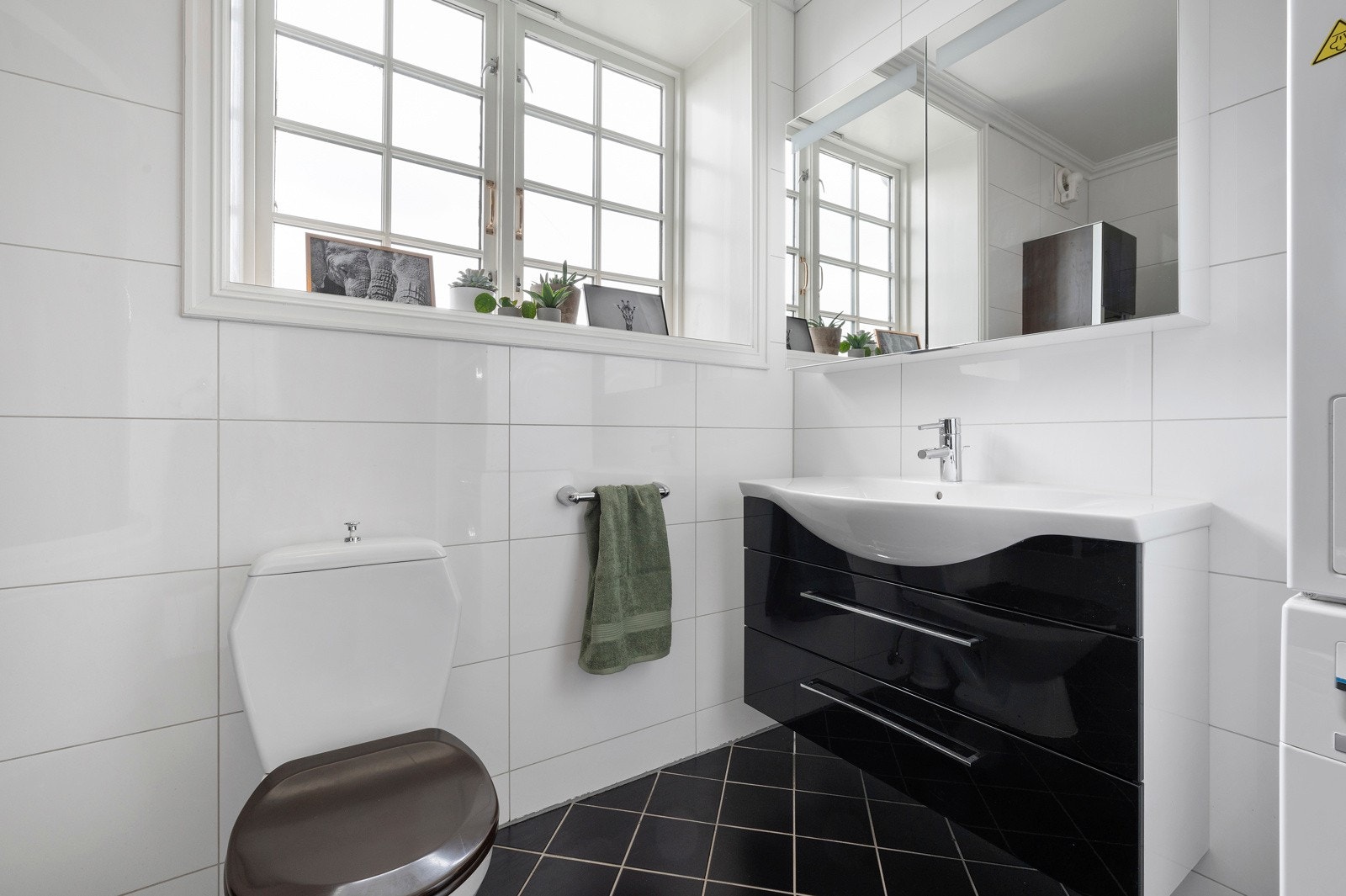 Det ene badet har sorte gulvfliser, hvite veggfliser, samt opplegg for vaskemaskin og tørketrommel i søyleløsning. Rommet er utstyrt med et kombinert badekar og dusjkabinett. Galleribilde