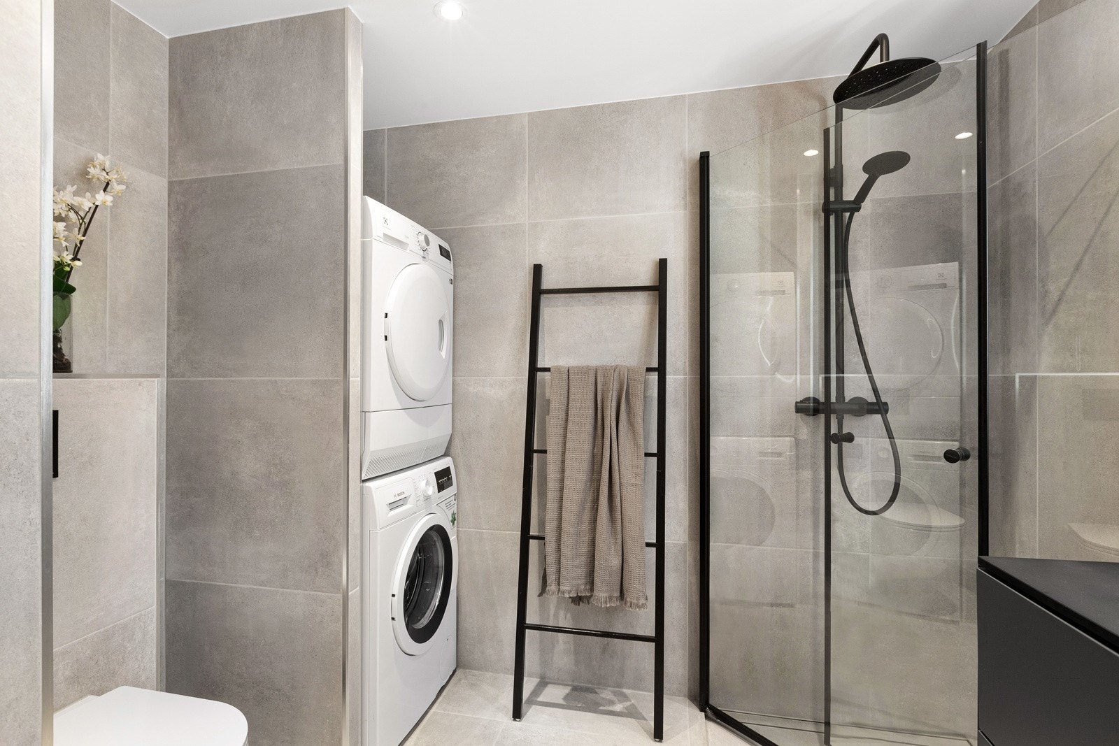 Badet har store,grå betongaktige fliser på gulv og vegger, med sorte detaljer på dusjdører, garnityr og blandebatteri. Galleribilde