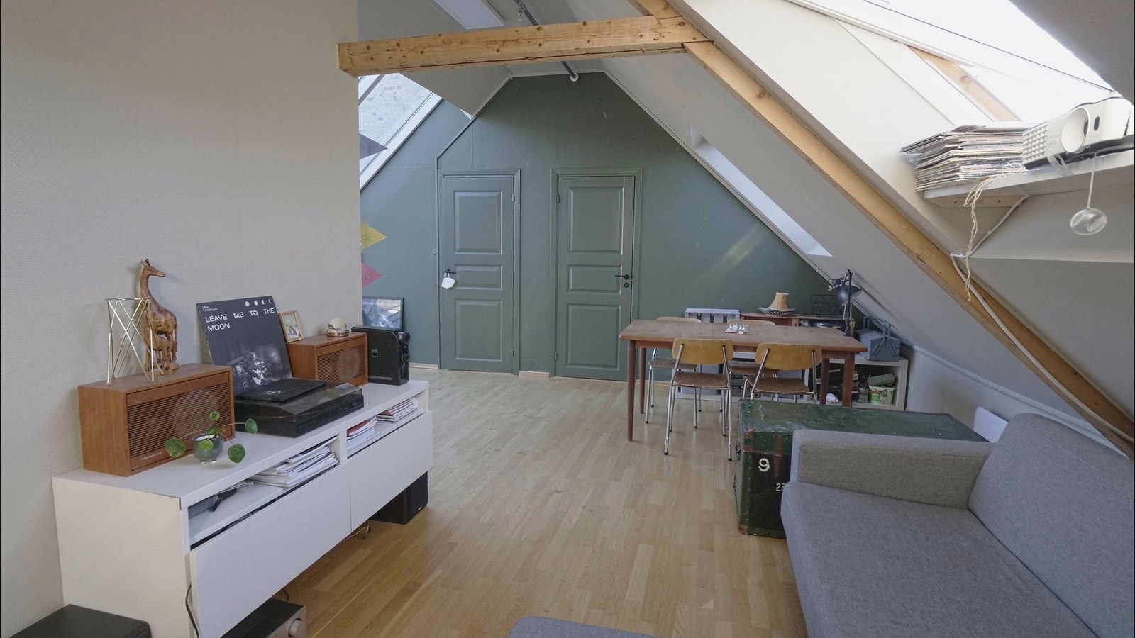 Kveldssol kommer gjennom skråvinduer. Møblert stue med sofa, prosjektor, lydanlegg, stuebord m. stoler, kaffebord. Galleribilde