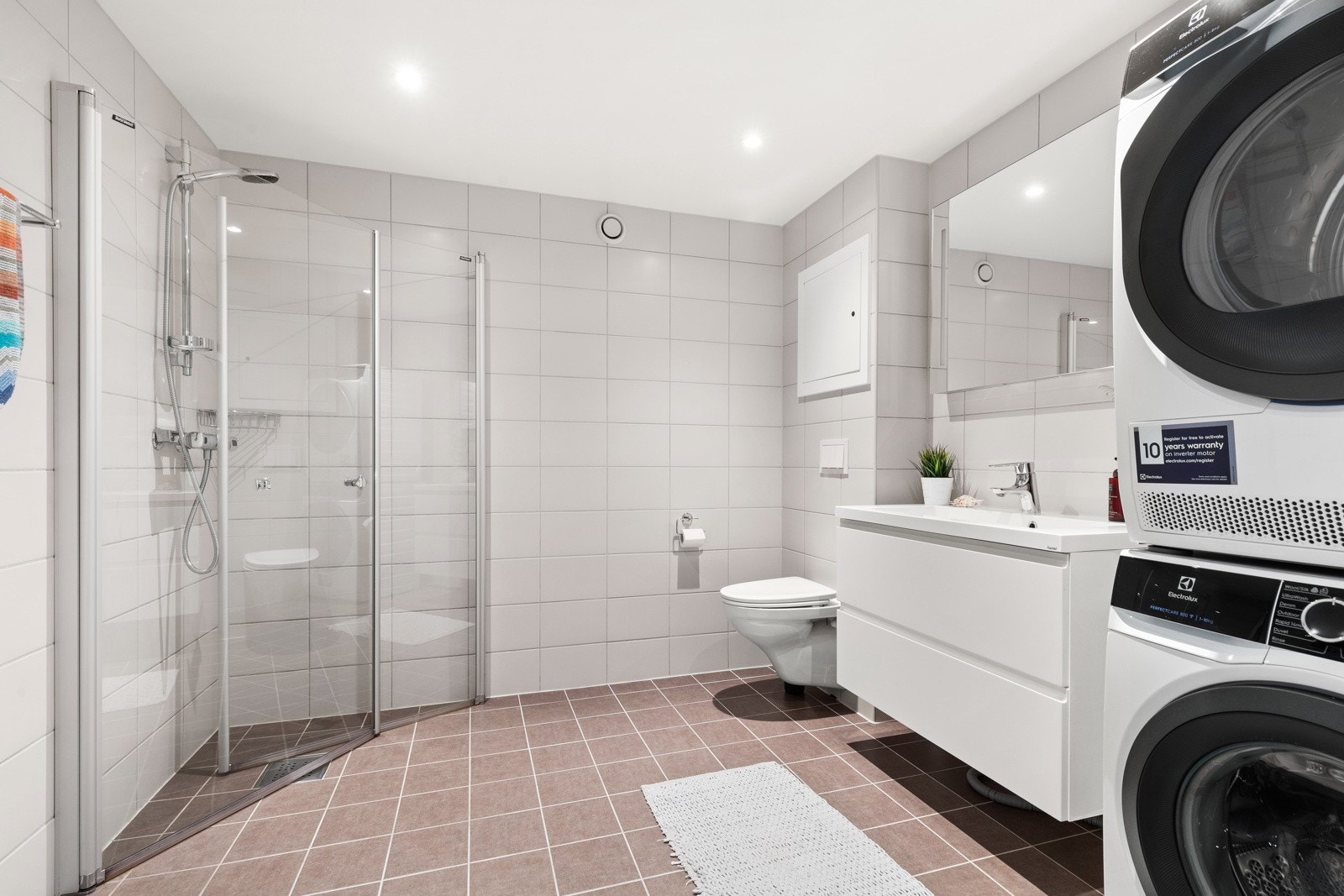 Badet er utstyrt med dusj med innfellbare glassvegger, servantseksjon med skuffer og heldekkende porselensvask, speil med lys, høyskap, vegghengt wc og opplegg for vaskemaskin/tørketrommel Galleribilde