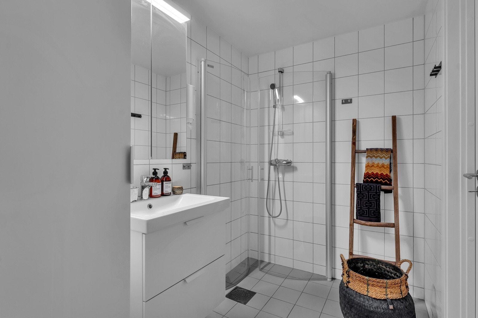 Badet er pent og lyst med grå gulvfliser, hvite veggfliser og gulvvarme. Det er opplegg for både vaskemaskin og tørketrommel. Innredningen består av en heldekkende servant på hvit skuffeseksjon, speilskap med overlys, et veggmontert toalett. Galleribilde