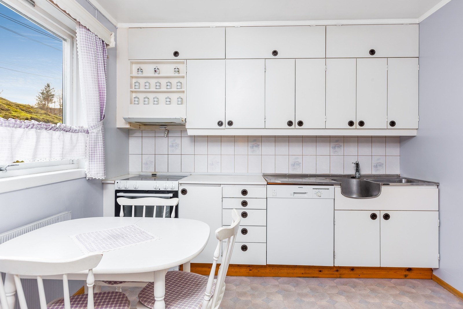 Kjøkkenet har eldre innredning av plater med overskap, benkeskap og skuffer, og foliert benkeplate. Vaskebeslag med oppvaskkum og utslagsvask. Ventilator. Galleribilde