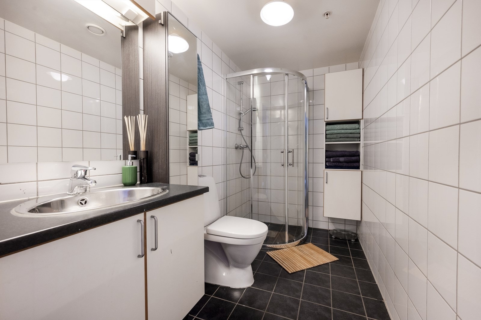 Badet er innredet med dusj, toalett, servant med vask og skuffeseksjon og speil med lys over. Galleribilde