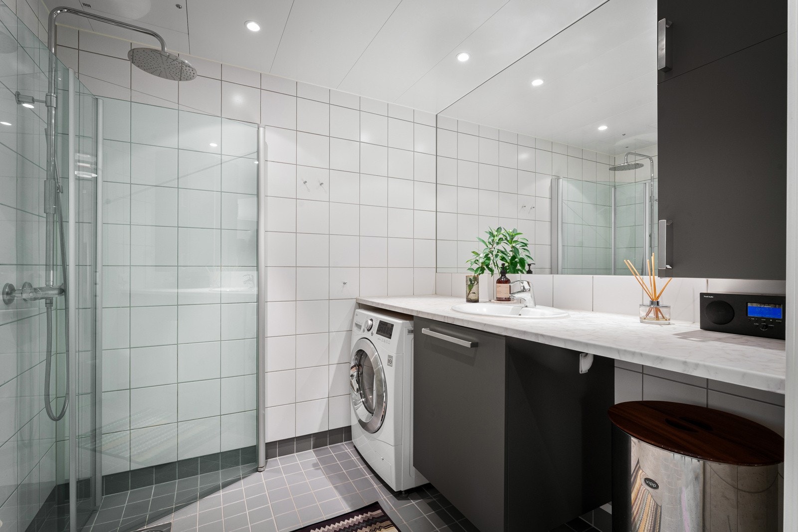 Baderommet har en klassisk stil med hvite veggfliser og grå gulvfliser over gulvvarme. Baderomsinnredningen består av over- og underskap med glatte, grå fronter og et stort speil med belysning. Galleribilde