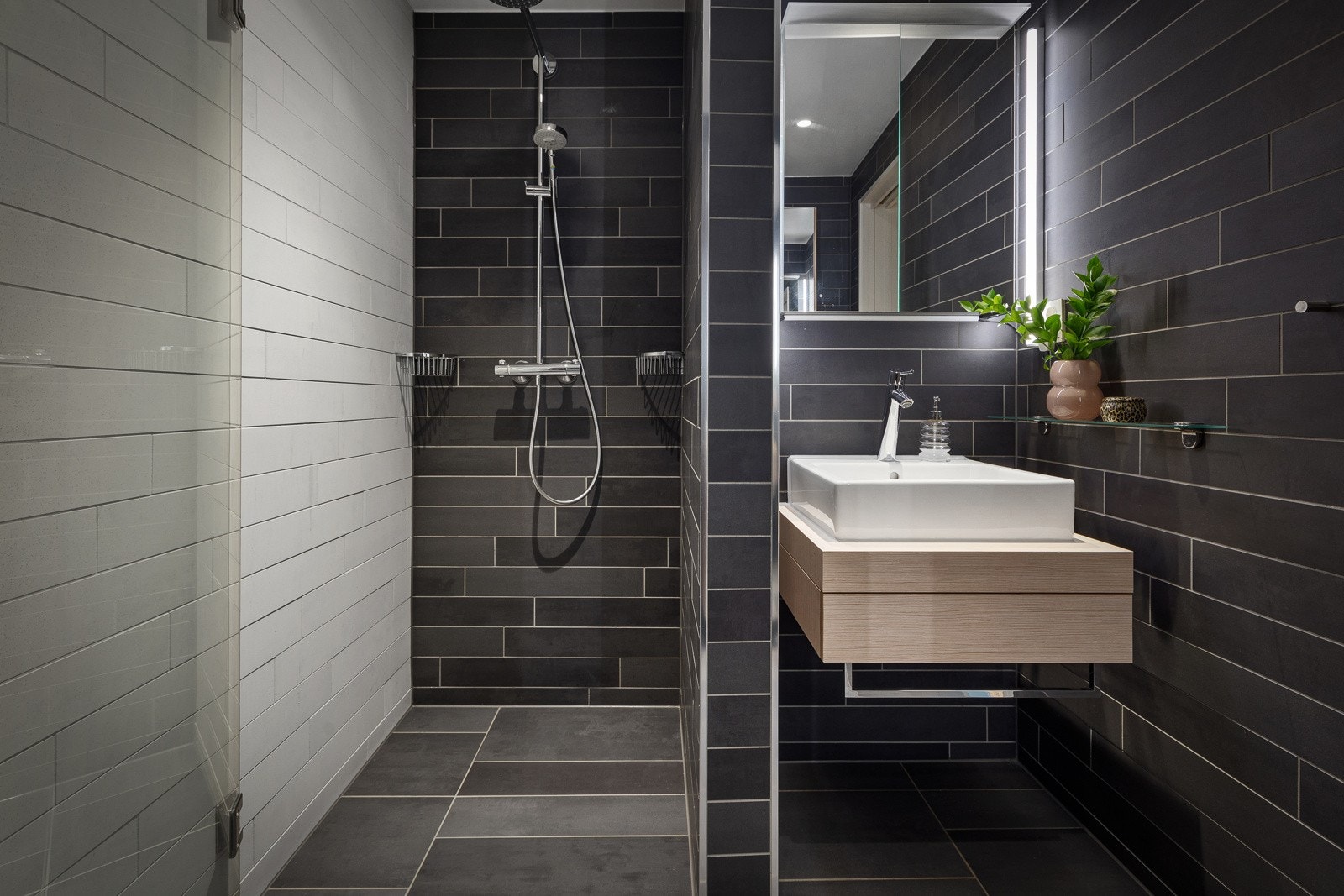 Badet ligger strategisk til ved entréen, og har en lekker kombinasjon av hvite og mørke fliser. Videre har badet dusjnisje med glassdør, vegghengt toalett, speilskap med belysning og et stilrent servantmøbel. Galleribilde
