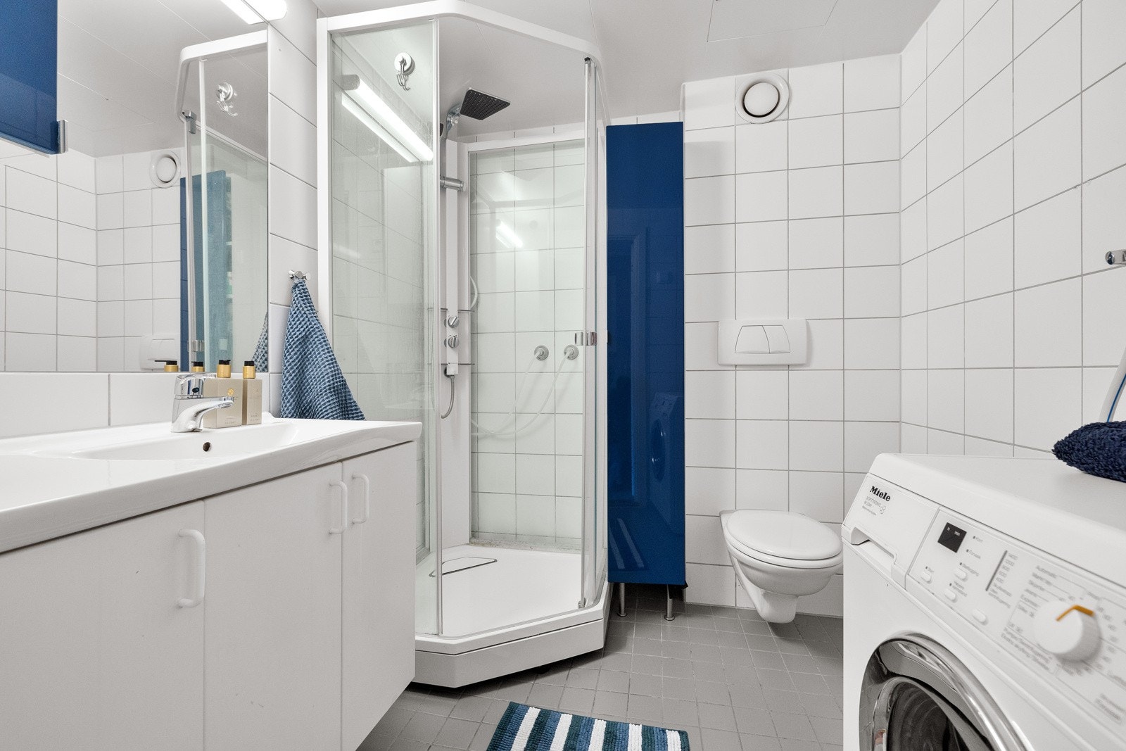 Flislagt bad som inneholder dusjkabinett, veggmontert wc, samt opplegg for vaskemaskin. Galleribilde