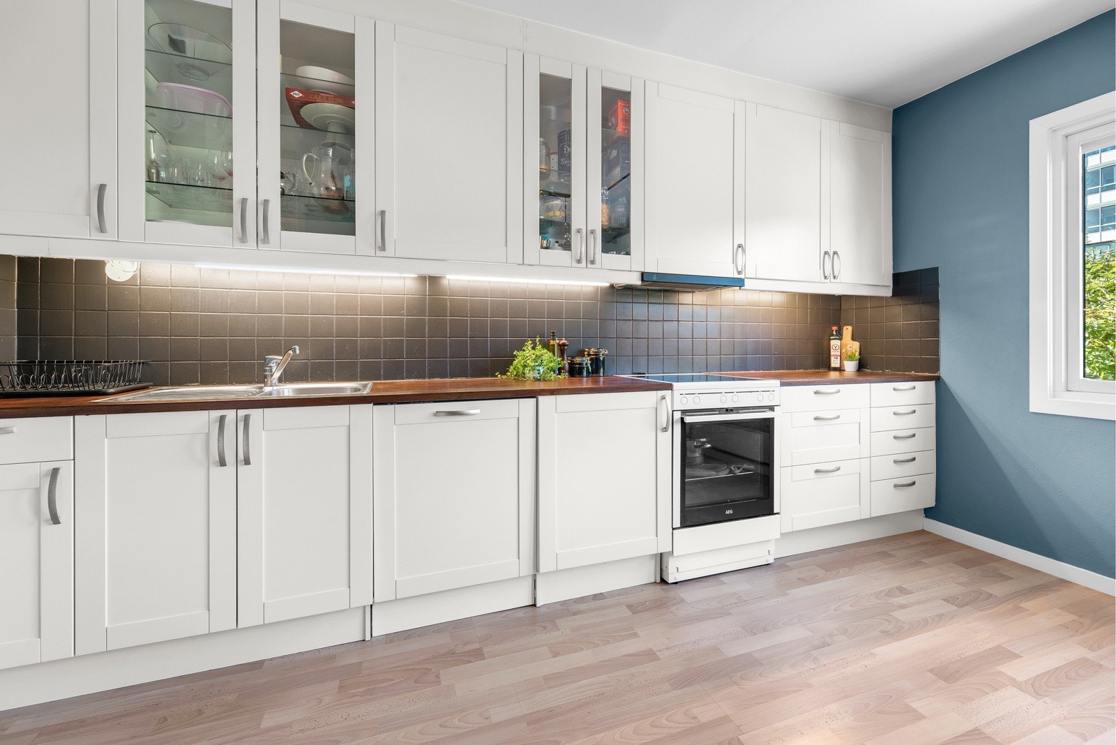 Hvit, profilert kjøkkeninnredning med vitrineskap og takhøy innredning, heltre benkeplate og kum i rustfritt stål. Her er det godt med oppbevaringsplass. Galleribilde