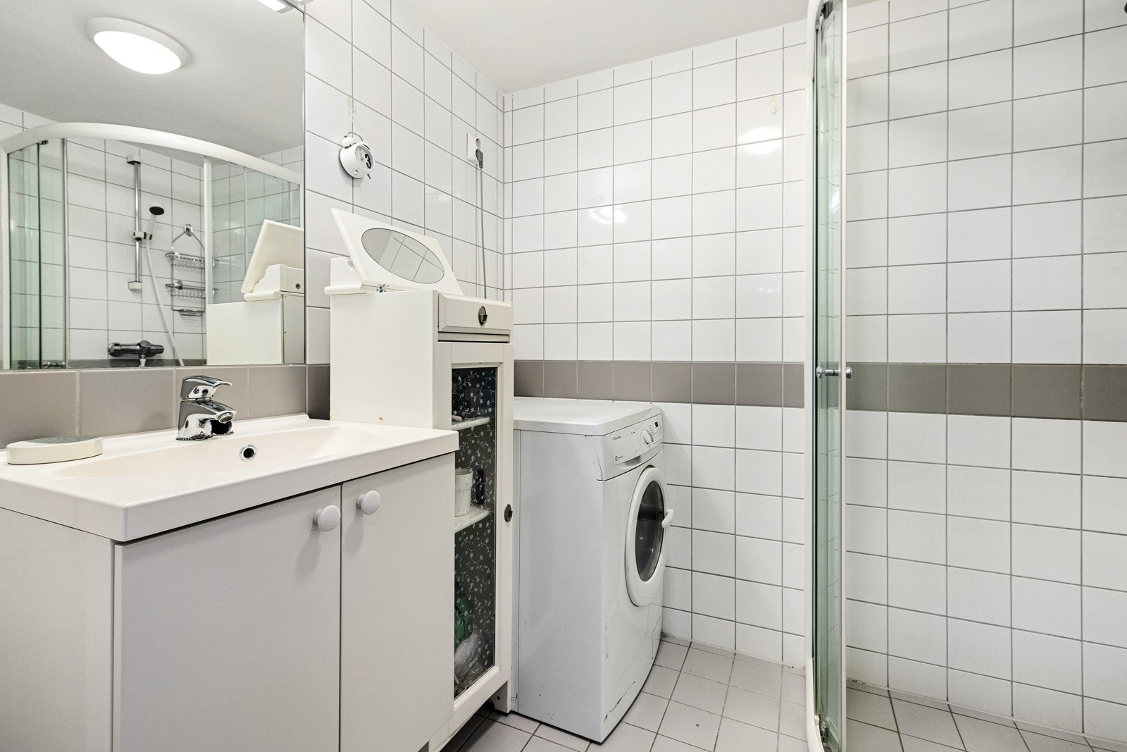 Baderom i hybeldel med dusjhjørne, toalett, servantskap, heldekkende servant, speil og opplegg for vaskemaskin og tørketrommel. Galleribilde