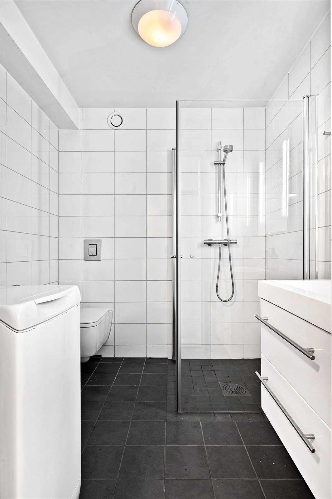 Flislagt bad med varmekabler i gulv. Vegghengt wc, dusjsone og servant i hvit skuffeseksjon. Opplegg for vaskemaskin. Galleribilde