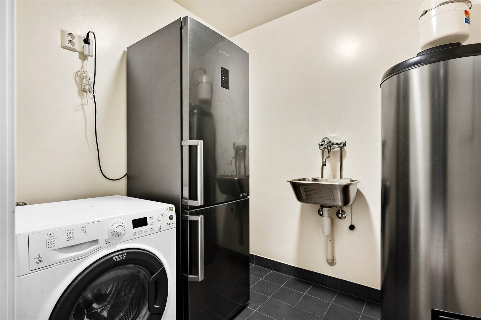 Praktisk vaskerom med flislagt gulv. Innredningen består av utslagsvask, varmtsvannstank og opplegg for vaskemaskin og tørketrommel. Galleribilde