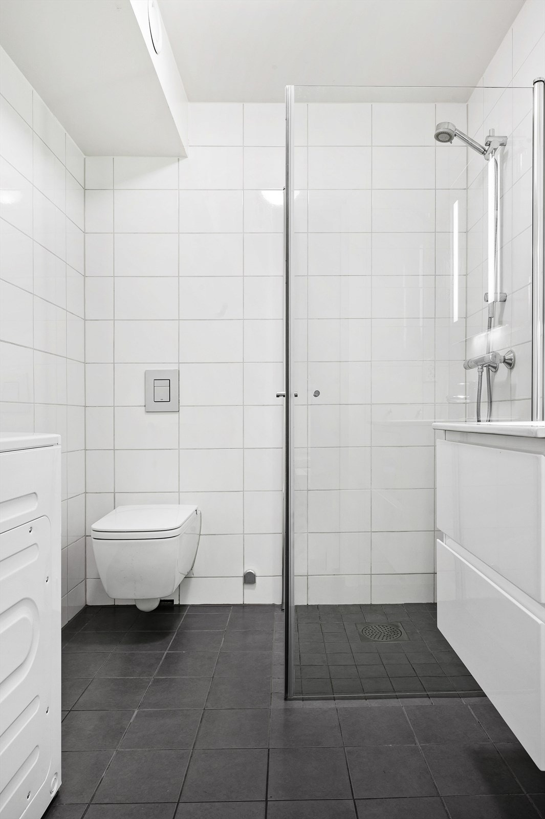 Flislagt bad med varmekabler i gulv. Vegghengt wc, dusjsone og servant i hvit skuffeseksjon. Opplegg for vaskemaskin. Galleribilde