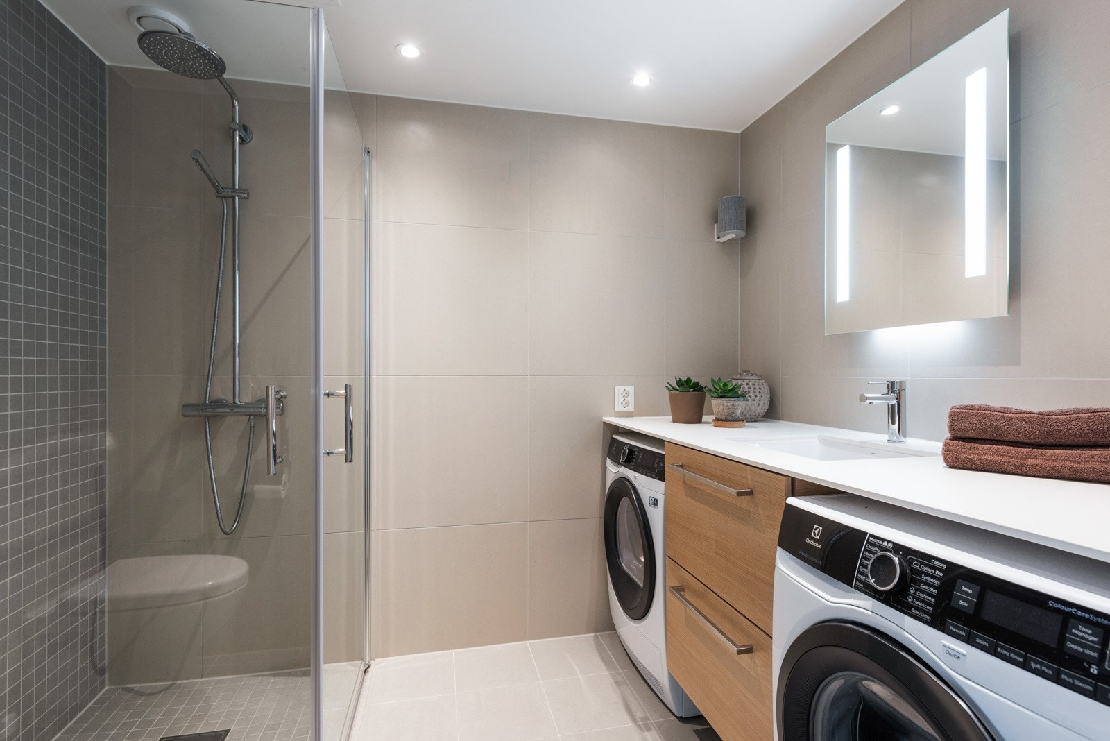 Stor baderomsbenk med opplegg for vaskemaskin og tørketrommel. Stor dusj i hjørnet bak svingbare glassvegger Galleribilde