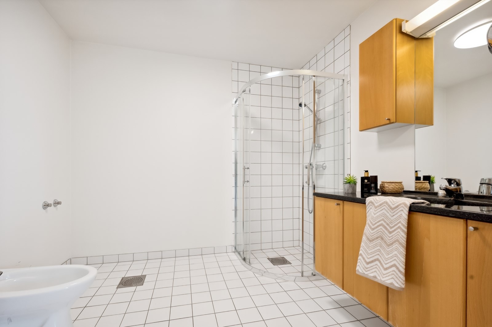 Badet er fra byggeår med malte overflater, fliser i dusjsone og fliselagt gulv med elektriske varmekabler. Det er innredet med speil, baderomsinnredning med servant, dusj med glassdører og toalett. Galleribilde