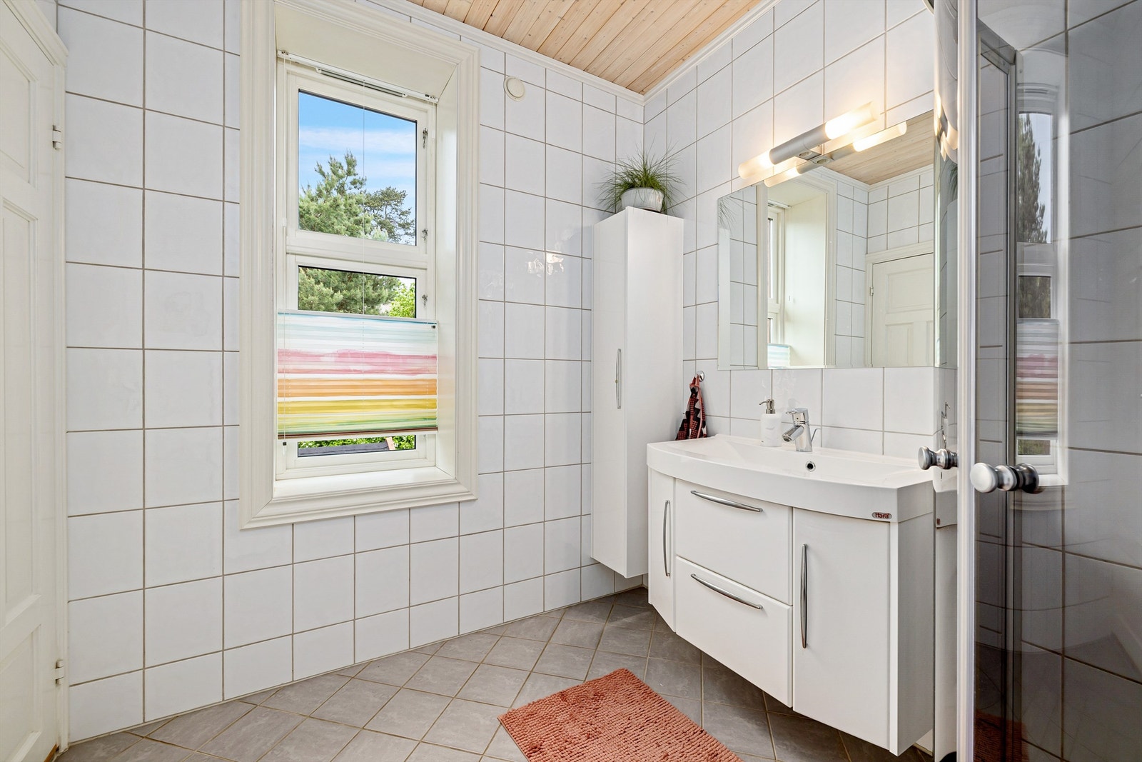 Badet har flislagt gulv med elektrisk gulvvarme og flislagte vegger. Himlingsflater i trepanel. Vegghengt servantskap med heldekkende servant og ett-greps armatur. Speil med overlys på vegg over servant. Høyskap. Dusjnisje med glassdører og dusjarmatur Galleribilde