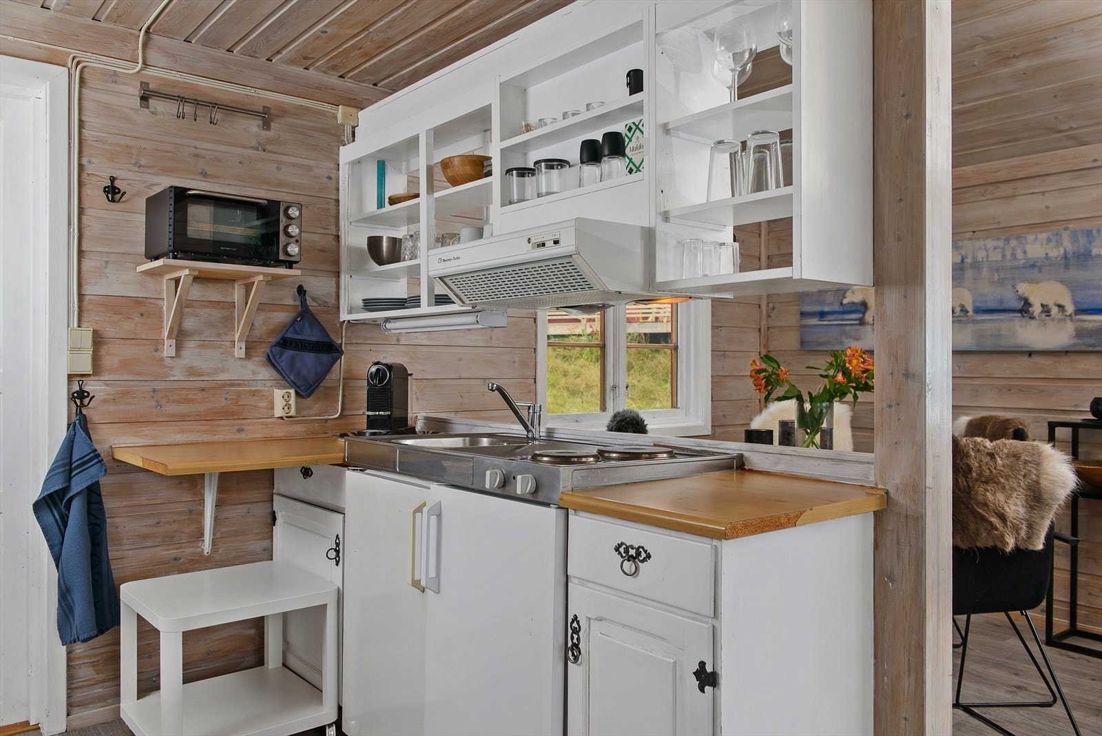 Enkelt kjøkken med lyse fronter og hyller over benk for oppbevaring. Kjøleskap og kokeplate. Galleribilde