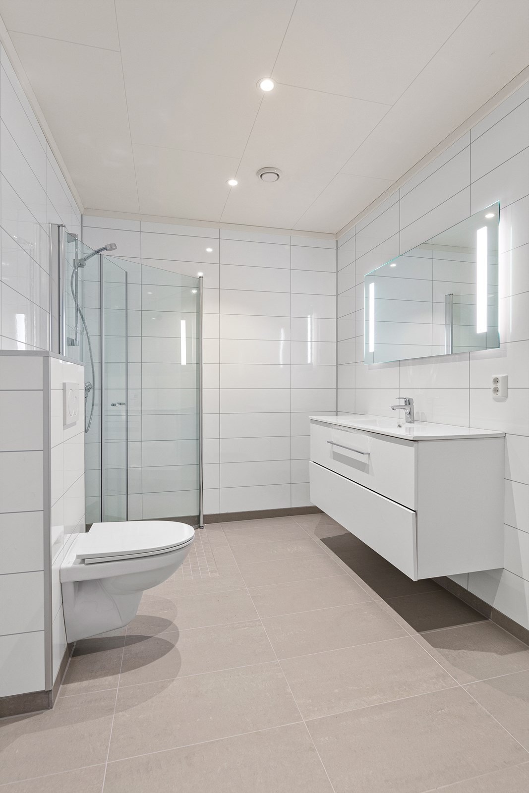 Badet er innredet med dusjhjørne med innfellbare glassdører, pen baderomsinnredning med speil og belysning og vegghengt toalett. Bildene er fra tilsvarende bolig, men speilvendt. Bildene er kun ment som illustrasjon av materialer og avvik vil forekomme. Galleribilde