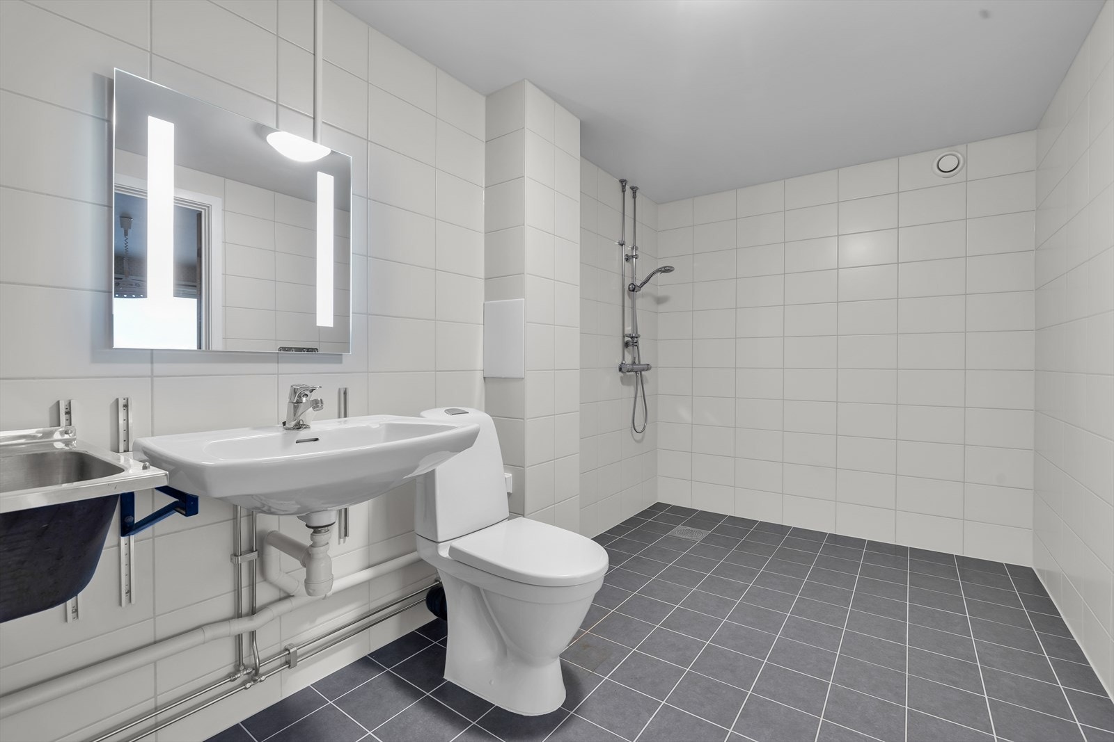 Romslig flislagt baderom bestående av servant, gulvstående toalett og dusjhjørne Galleribilde