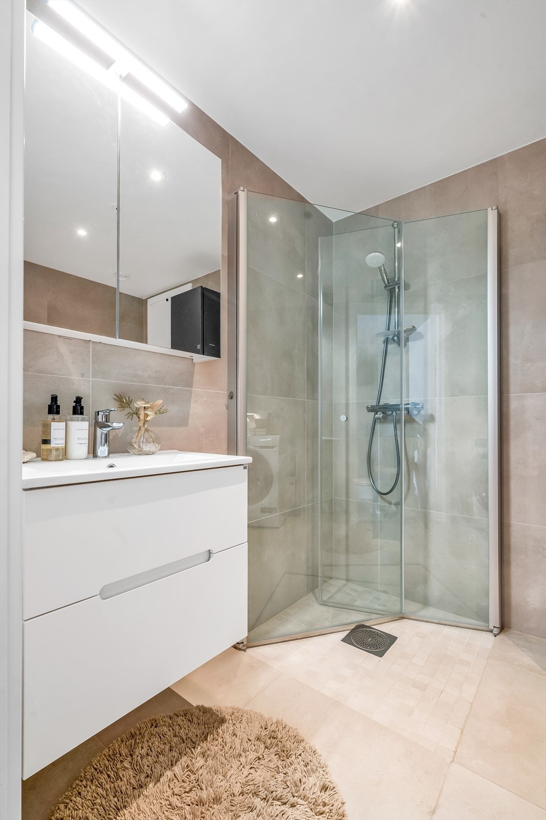 Badet er utstyrt med vegghengt toalett, dusjhjørne med innfellbare glassdører, servant med skuffeinnredning og speilskap med belysning Galleribilde