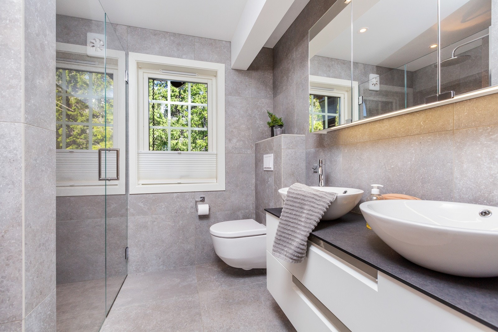 Moderne innredning med to runde vasker på benkeplate, speilskap, vegghengt toalett og dusjnisje med glassdører. Galleribilde