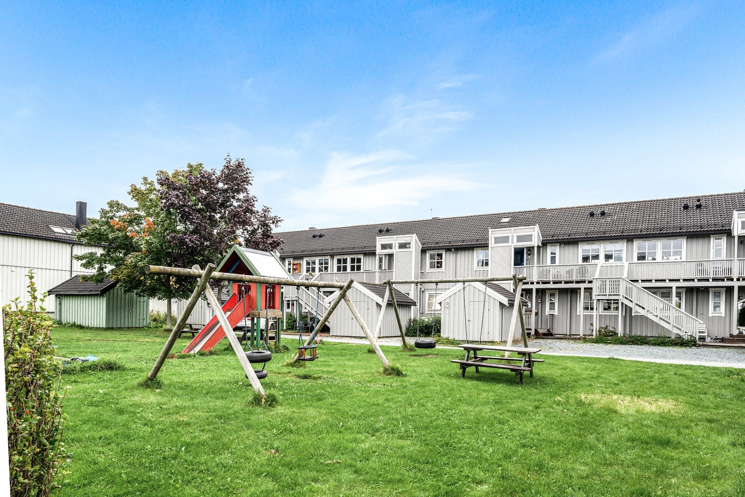 Sommerbilde av lekeplass og fasade. Fasade ble malt og beiset i 2018/19. Sportsbod rett nedenfor inngangsparti. Galleribilde