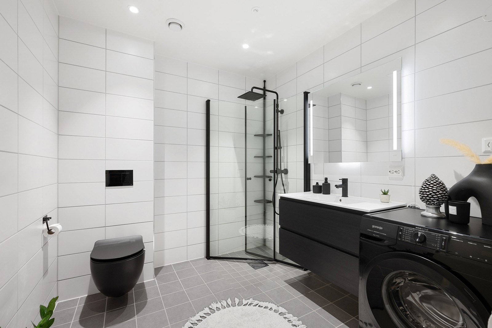 Badet er lekkert i moderne stil med sorte detaljer, inkludert sort, veggmontert toalett, opplegg for vaskemaskin, samt et dusjhjørne med innfellbare glassdører og sort dusjgarnityr med både hånddusj og regnfallsdusj. Galleribilde