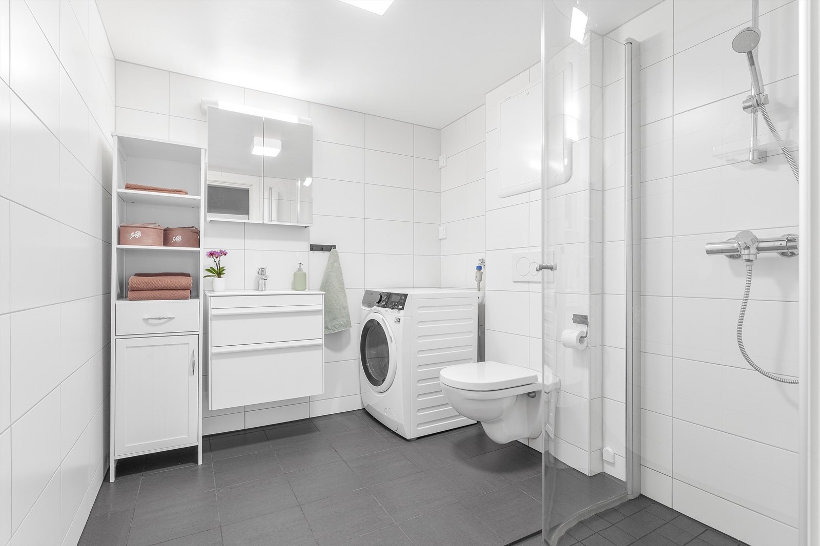 Moderne, flislagt bad fra 2018. Innredningen består av vegghengt wc, dusjsone med buede, innfellbare dusjvegger, samt servant i hvit skuffeseksjon med speilskap og belysning over. Galleribilde