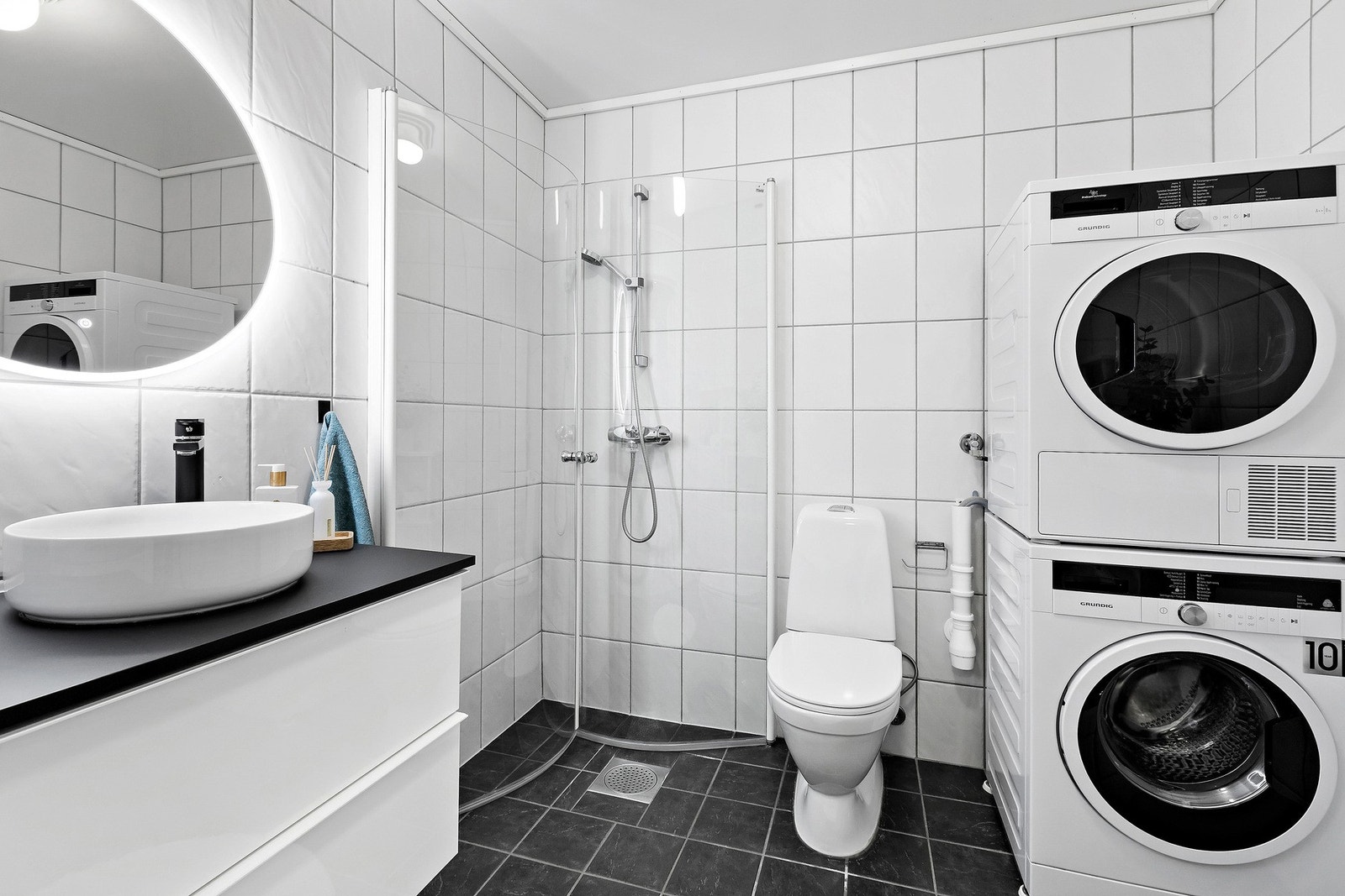 Delikat flislagt baderom med infellbare dusjvegger i glass, opplegg for vaskemaskin og tørketrommel, wc og moderne innredning. Galleribilde