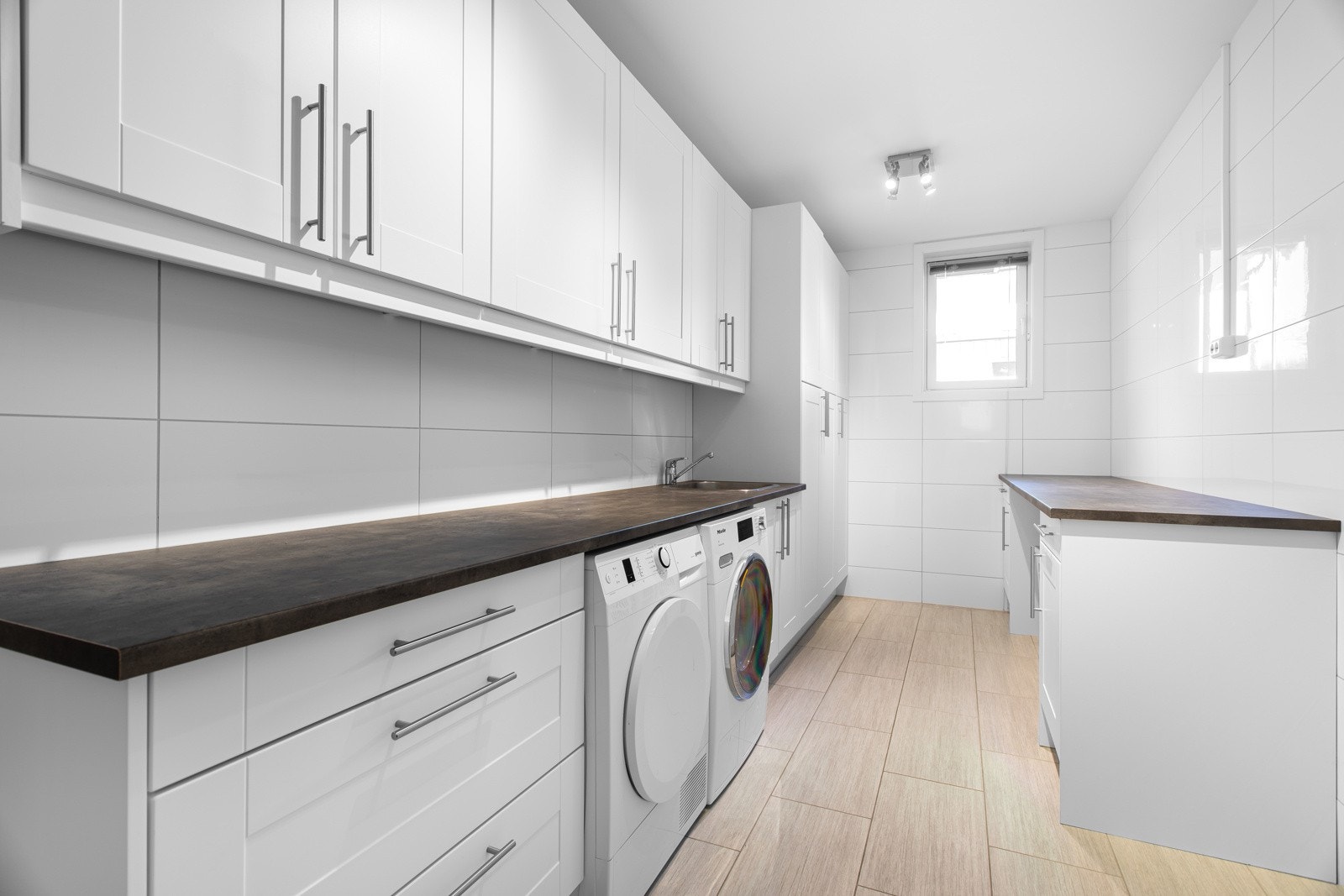 Rommet er lyst og delikat med flislagte gulv og vegger. Innredet med arbeidsbenk med vask og overskaper. Under arbeidsbenken er det i tillegg til skuffer og skap, plass for vaskemaskin og tørketrommel. Klessjakt fra etasjene over. Galleribilde