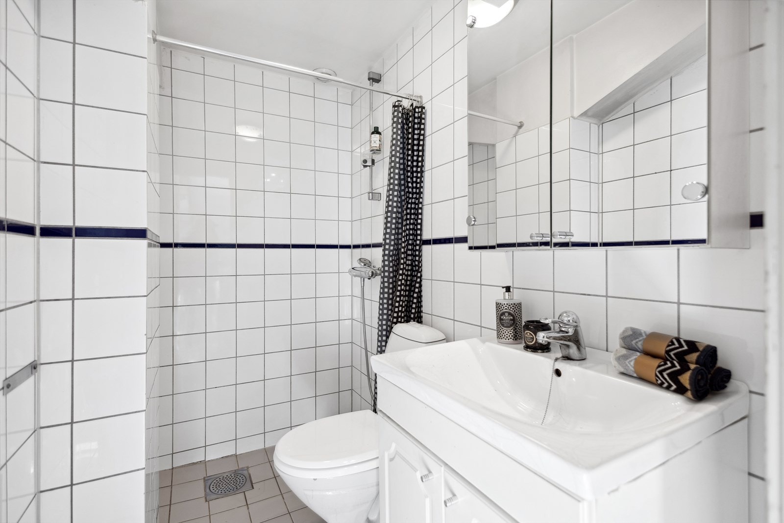 Badet har tidløse, hvite veggfliser og grå fliser på gulv med gulvvarme. Dusjnisje med forheng, stående klosett og helservant på baderomsskap. Speilskap med belysning over servant. Galleribilde
