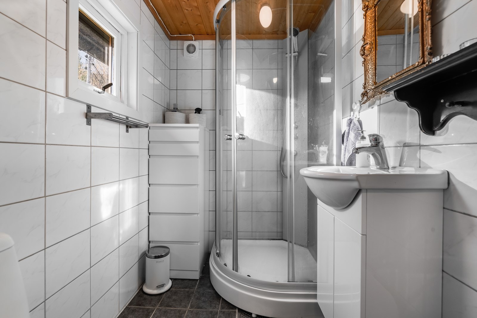 Her er det flis på gulv og vegger, og badet inneholder toalett, dusjkabinett og innredning med heldekkende hvit vask og underskap med slette fronter. Det er mekanisk avtrekk. Galleribilde
