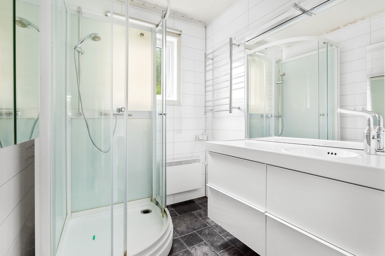 Badet i første etasje ble pusset opp i 2012. Rommet har mørkt belegg på gulv med flismønster og hvite flisplater på vegg. Videre består innredningen av speil med belysning, enkel servant på hvitt servantmøbel og dusjkabinett. Vurdert som TG 2 av takstmann Galleribilde